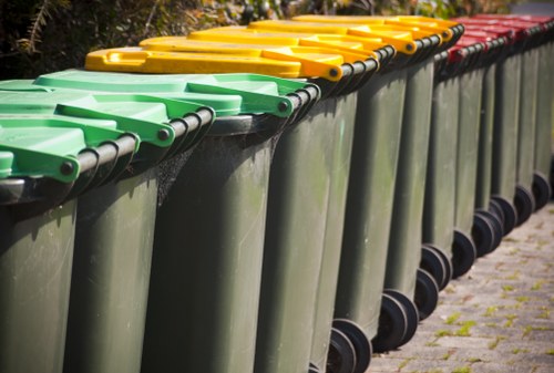 A row of large green wheelie bins lined up along a paved outdoor area, with yellow and red lids on the top of some bins. The bins are positioned on the right side of the image, with their wheels visible at the bottom. The lids are closed and clean, showing a slight sheen in the sunlight, and the background includes trees and bushes. The scene reflects a typical waste collection point in a residential or commercial area near Charlton, where Office Clearance Charlton offers rubbish removal services. The ground consists of asphalt with some small patches of moss or grass growing between cracks, indicating an outdoor environment dedicated to waste disposal or rubbish collection, consistent with local rubbish management practices in London or the surrounding postcode area.