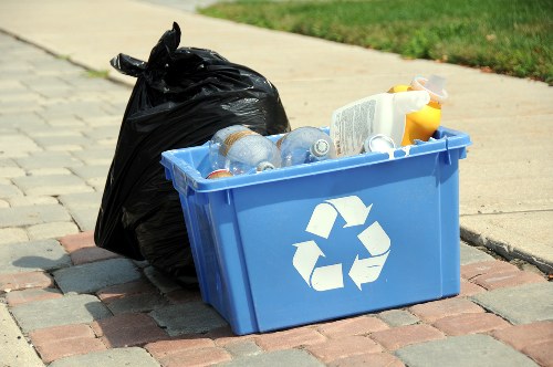 On a paved sidewalk with interlocking brick pattern in Charlton, a blue plastic recycling bin featuring the universal recycling symbol is filled with clear plastic bottles, a white container, and a yellow bottle, indicating mixed recyclables. Beside the bin, a black plastic rubbish bag is tied at the top, appears partially filled, and is leaning slightly against the bin. The background shows a strip of well-maintained green grass, suggesting the rubbish removal service by Office Clearance Charlton is staged for collection or disposal in the local area. The scene depicts typical waste management collection preparations, with the items placed on the pavement surrounding the bin and bag, ready for pickup or transportation to appropriate disposal or recycling facilities within the vicinity.