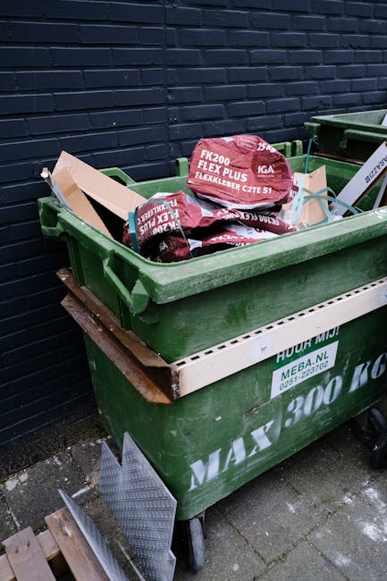 A worker wearing a high-visibility yellow and red uniform is operating a large, red rubbish collection vehicle on a street in Charlton. The back of the vehicle is open, revealing mechanical lifting co