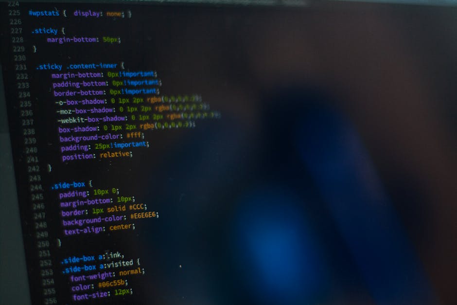 Close-up view of a computer screen displaying lines of colourful source code in green, orange, blue, and purple text against a dark background. The text appears slightly blurred, with some sections sh